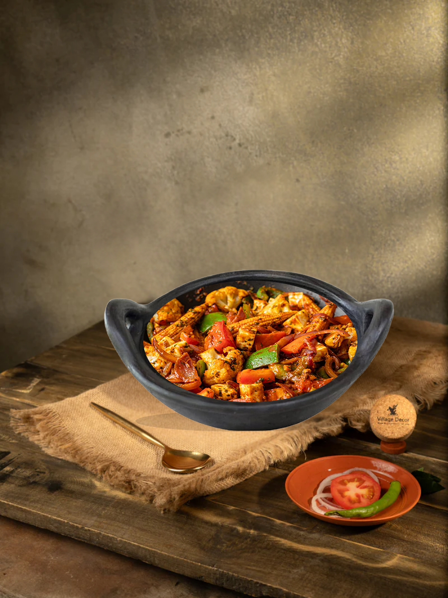 Traditional earthen black clay kadai filled with sautéed mixed vegetables, placed on a rustic wooden table with brass spoon and garnishing plate on the side.