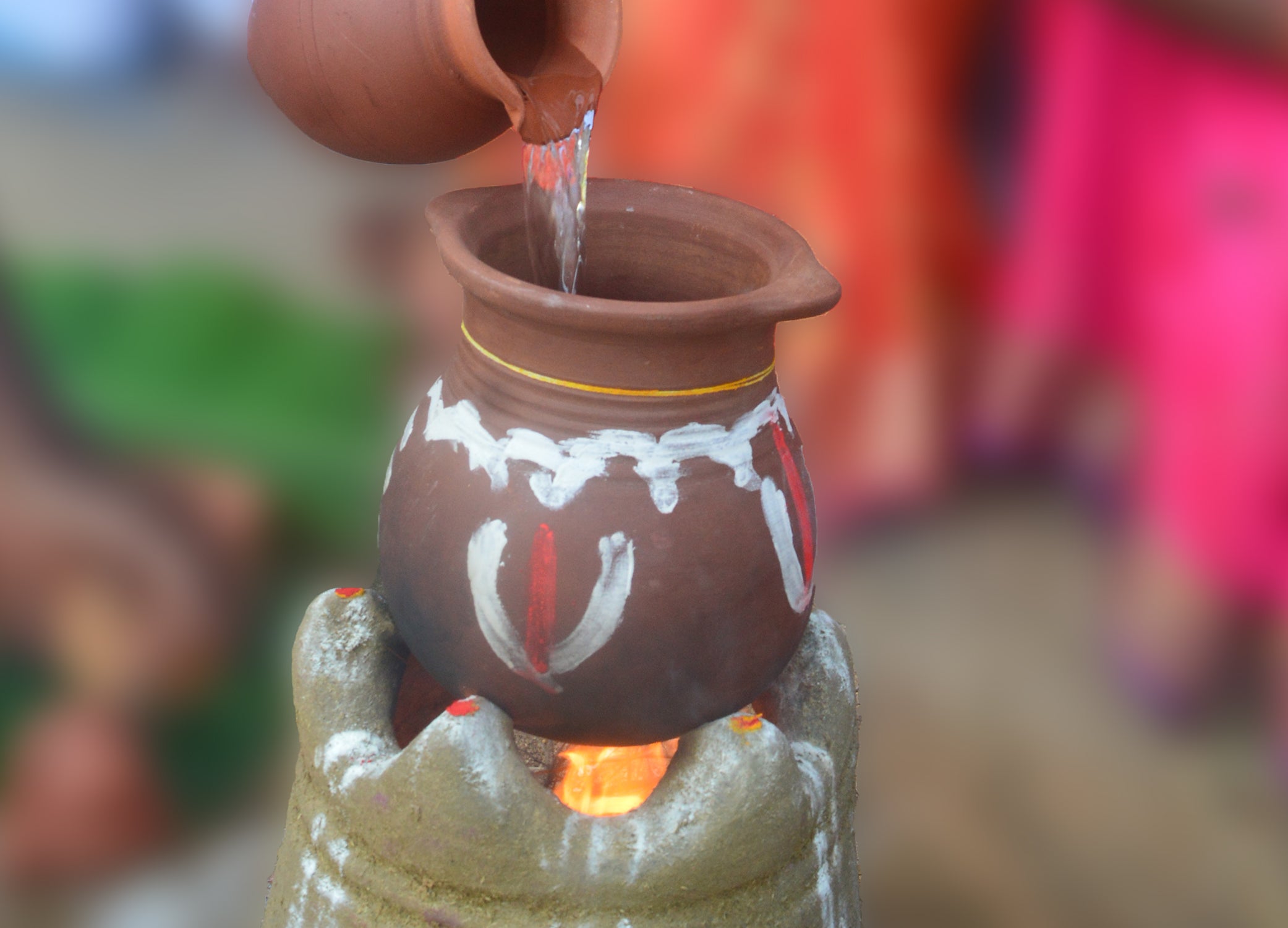 Water being poured into a decorated earthen clay pongal pot placed over a traditional wood-fired stove during Pongal festival preparation.