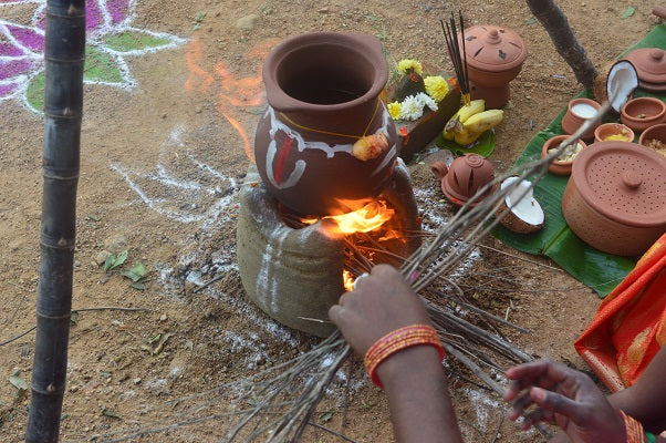 Traditional Pongal celebration showing an earthen clay pot on firewood, decorated with turmeric, flowers, and surrounded by banana leaves, brass utensils, and festive items.