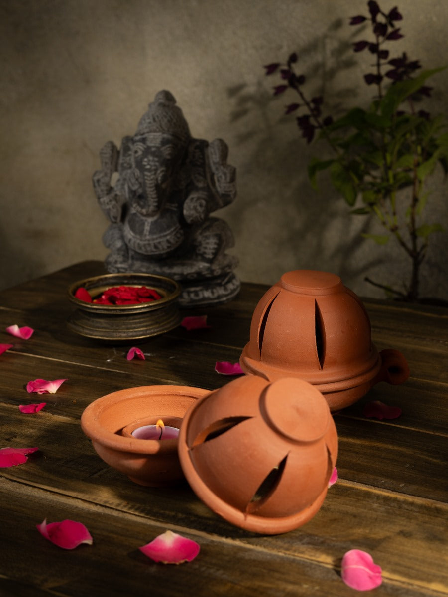 Handcrafted terracotta diya with lid placed on wooden surface, surrounded by rose petals, with a Ganesha idol and tulsi plant in the background, perfect for pooja or festive decor.
