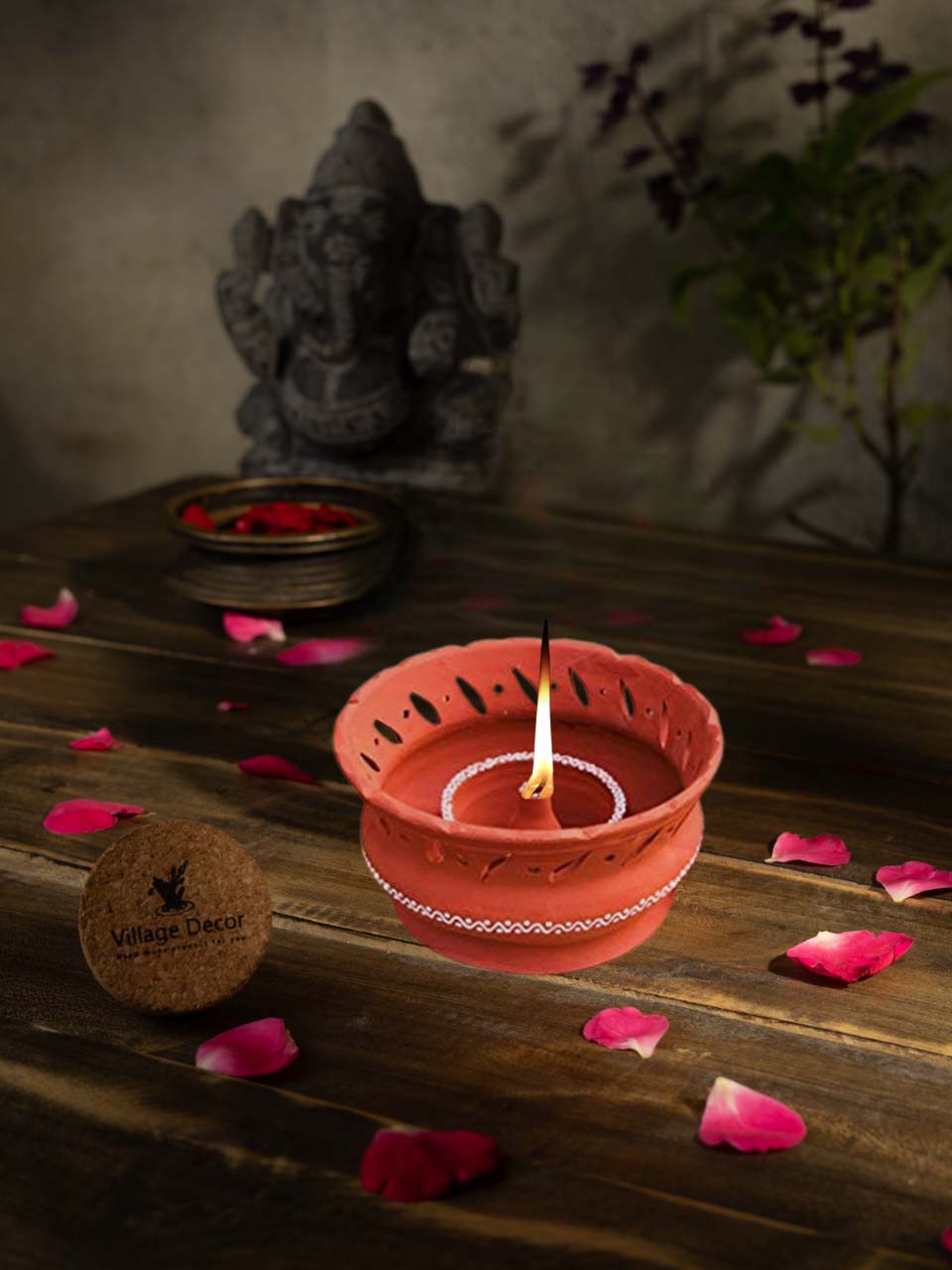 Lifestyle view of a terracotta vallar diya with intricate cut-out design and painted detailing, placed among rose petals with a burning flame in the center.