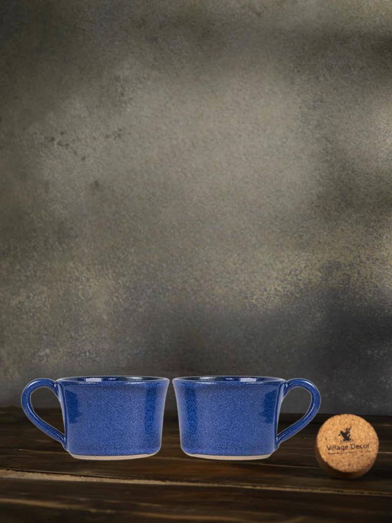 Two blue ceramic mugs on a wooden surface with a cork coaster and wooden block against a dark gray background.