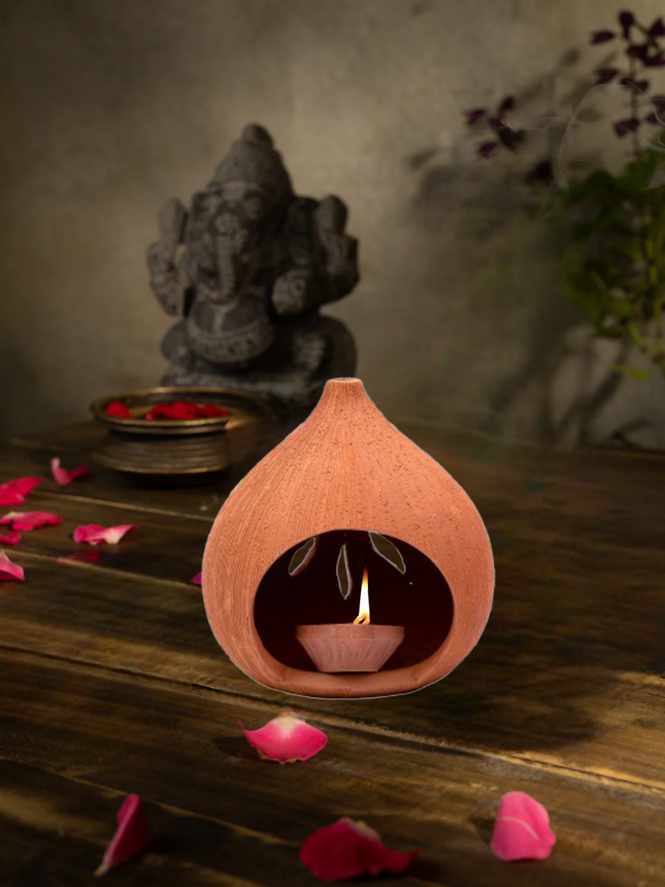 Terracotta oil lamp with flame on a wooden surface with pink petals and a stone statue in the background.