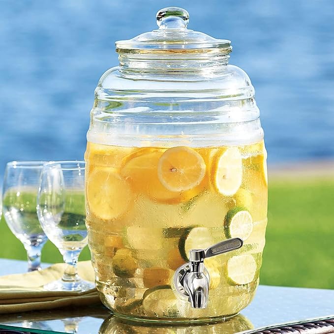 Glass jar beverage dispenser filled with lemon and lime drink, featuring a stainless steel tap, placed outdoors near a lake with glasses beside it.

