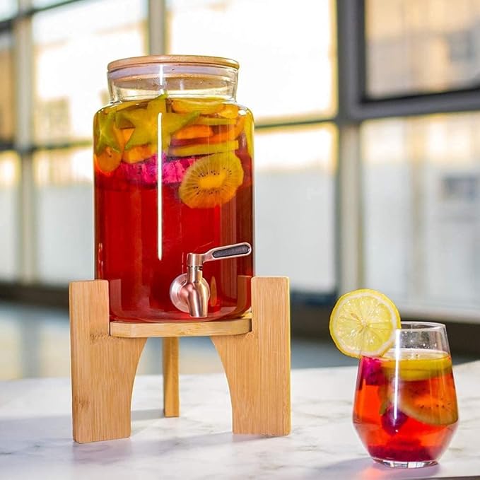 Glass beverage dispenser filled with fruit-infused drink, placed on a wooden stand with a stainless steel tap, next to a glass of colorful summer refreshment.